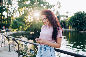 Cheerful young lady reading messages on smartphone in park