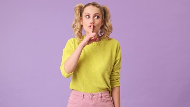 A cute blonde in a yellow sweater looks around with a smile and shows silence gesture in an isolated studio on a purple background