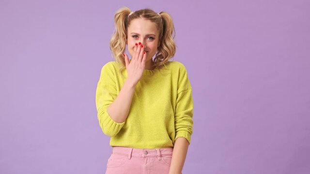 A cute blonde girl heard a funny joke and shyly laughs at her, covering her mouth with her hand in an isolated studio on a purple background