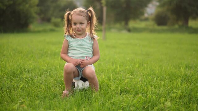 Childhood, games and entertainment, sports, physical culture concept - little girls in shorts and two ponytails play sitting on soccer black and white ball on bright green lawn in park at sunset