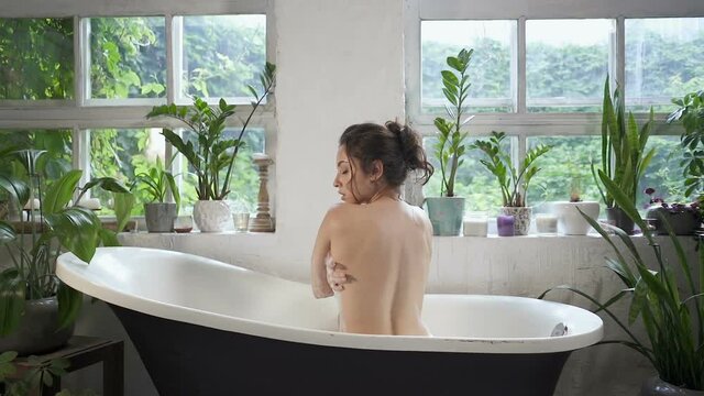Attractive brunette woman sitting in the bathtub and looking at the camera. Slowmotion.