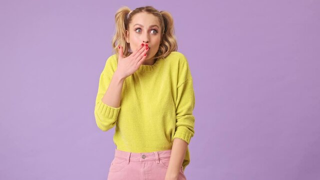 A fine blonde girl with surprise and smile covers her mouth with her hand in an isolated studio on a purple background