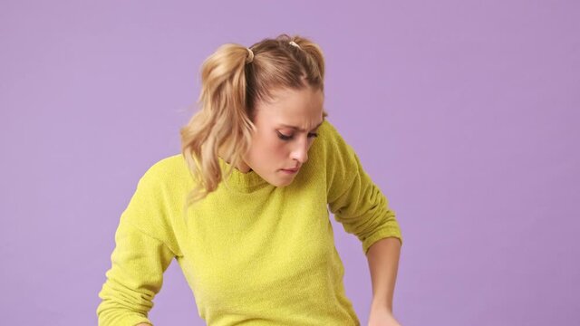 A pretty forgetful blonde in a yellow sweater is trying to find something in her pockets, but can not do it in an isolated studio on a purple background