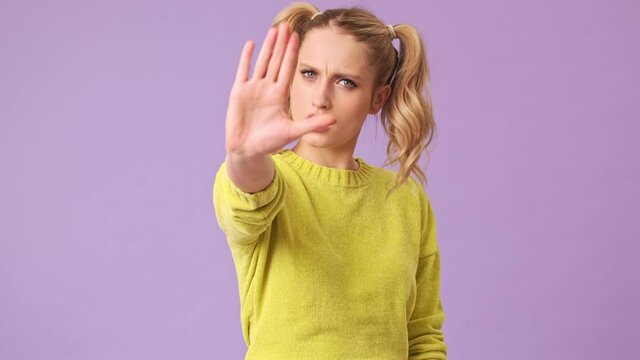 A beautiful blonde girl maliciously looks at the camera and demands to stop talking, showing with her hand, shaking her head in the negative in an isolated studio on a purple background