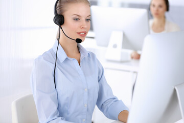 Call center office. Beautiful blonde woman using computer and headset for consulting clients online. Group of operators working as customer service occupation. Business people concept