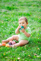 Cute kid in a green bodysuit sitting on the grass in summer plays with developing toys. The baby gnaws silicone cubes. Child development up to one year