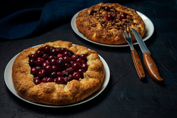 Two galettes with ripe red cherry and walnut filling on dark blue background. Homemade sweet open pie with cutlery.