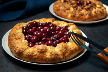 Two galettes with ripe red cherry and walnut filling on dark blue background. Homemade sweet open pie with cutlery. Close-up.