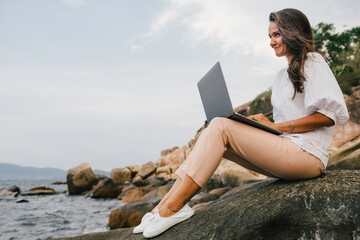 Young successful, smiling girl working using a laptop, sitting on a rocky seashore. traveling, blogger, freelancer, content plan, work online