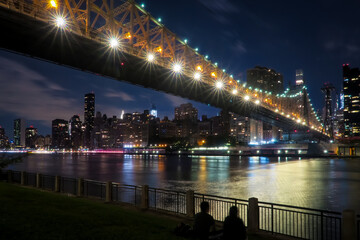 Fototapeta na wymiar New York, USA - September 8, 2019 - The Queensboro Bridge and East River. Manhattan skyline is in the background. A couple (silhouette) sitting by the river.