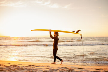 Young fit surfer in wetsuit with blank yellow surfing longboard walk from water at sunset ocean. Water sport adventure camp and extreme swim on summer vacation.
