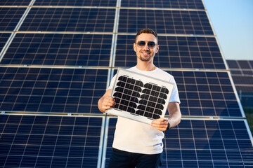 Smiling young man holding a small pv module looking at the camera, solar plant behind his back,...
