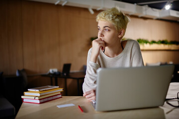 Attractive blonde female designer pondering on idea for project sitting in cafe with loft interior,skilled freelancer creating plan for completing task with marketing strategy using modern technology.