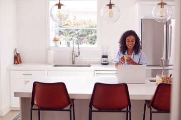 Woman Wearing Pyjamas Sitting In Kitchen Working From Home On Laptop