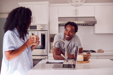 Fototapeta na wymiar Couple Wearing Pyjamas Standing In Kitchen Working From Home On Laptop