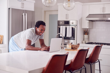 Man Wearing Pyjamas Standing In Kitchen Working From Home On Laptop
