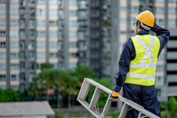 Asian maintenance worker man with safety helmet and green vest carrying aluminium step ladder at construction site. Civil engineering, Architecture builder and building service concepts © zephyr_p