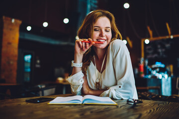 Beautiful young woman enjoying creative job in cafe interior solving problem with project in good mood, smiling entrepreneur checking notes in dairy making schedule of daily work holding pen