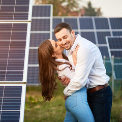 Close-up shot of a young couple standing between solar panels, woman is kissing a man in a cheek, bent backwards, man hugging her, bent forward