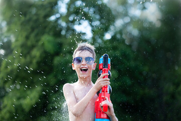 Smiling boy in glasses play with water gun at hot sunny summer day