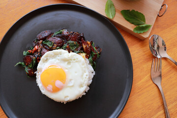Chinese sausage fried with basil and fried egg with rice in the black round dish, Cooking ingredients with spoon and fork.