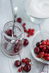 Currants and cherries in a glass jar, a glass plate with berries and sugar on the background of a table of white boards; light background for summer recipes for making homemade jam