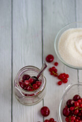 Currants and cherries in a glass jar, a glass plate with berries and sugar on the background of a table of white boards; light background for summer recipes for making homemade jam