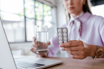 cropped view of woman holding glass of water and blister pack with pills near laptop