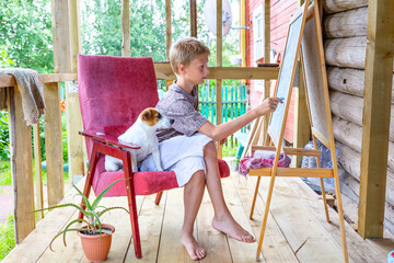 Caucasian blond-haired boy sits in a pink armchair and draws on an easel with crayons, plein air. Nearby sits a dog Jack Russell Terrier. Portrait of a teenager with a dog. Aloe, a houseplant in a pot