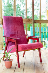 An old vintage armchair of dark pink color stands on an open veranda on a plank floor, next to a flower pot with aloe. Summer day, a cozy place to read, think, relax. Restoration of old furniture.