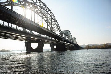 A long arched iron bridge over a wide river. A sun breaks through it.