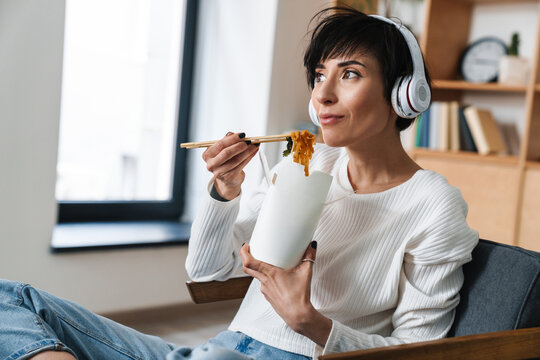 Image of pleased woman using headphones while eating asian noodles