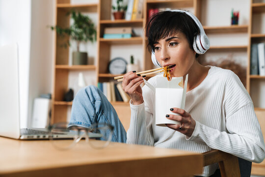 Image of serious woman eating asian noodles while working with laptop