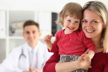 Portrait of happy young pretty woman with little baby visiting pediatrician in clinic with doctor on the background. Healthcare concept