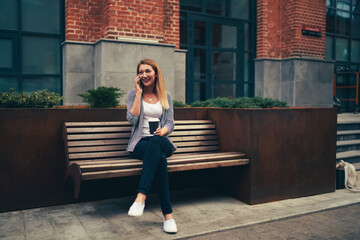 Smiling young attractive female student having mobile conversation with friends using application on smartphone.Happy blogger having fun while talking on telephone during free time at urban setting