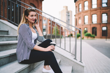 Fototapeta premium Young caucasian woman with nice smile talking on phone in pleasant atmosphere outdoors sitting on university stairs and having takeaway coffee, contemporary technology and communication concept