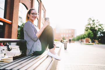 Smiling trendy caucasian woman 20s sitting on bench and holding cup of coffee outdoors at sunny day, portrait of cheerful stylish hipster girl looking at camera while enjoying summer weather