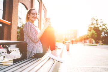 Smiling trendy caucasian woman 20s sitting on bench and holding cup of coffee outdoors at sunny day, portrait of cheerful stylish hipster girl looking at camera while enjoying summer weather