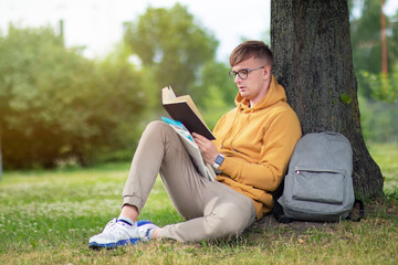 Handsome guy, man, young college or university student or pupil in glasses sitting outdoors in summer park reading book leaning on a tree, learning lessons, preparing for exam in university or college