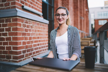 Portrait of successful female software developer looking at camera in break of distance job and working on laptop computer, smiling trendy copywriter spending time with netbook at street cafe