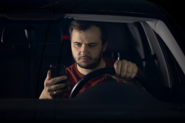 Unshaven confident serious young millenial male driver sits at steering wheel of car at night tired looking at smartphone worried concerned about problems. safe driving concept. Loneliness Concept.
