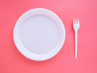 White plastic plate and fork on pink background, flat lay, top view. Bright minimalistic concept of eating and table setting. Bright slide to your presentation. The problem of disposal of plastic