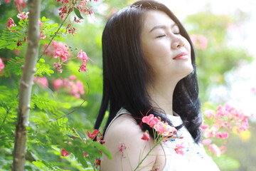 Naklejka premium Beautiful asian woman in white dress close her eyes and smelling in blooming flowers with natural in garden sunlight. Thai or Chinese Girl enjoy on holiday