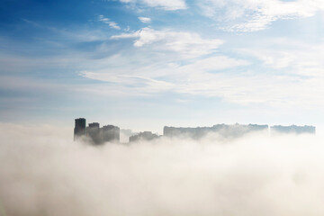 Obraz premium White fog in the city, residential buildings in mist against the blue sky with clouds. Urban landscape in early morning, beautiful background for wet weather