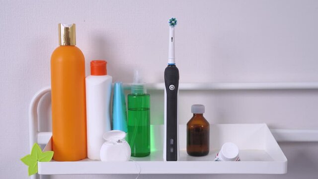 On shelf in bathroom are dental floss in box, toothpaste and tubes with various hygiene products, care and cleansing of face, body, hair and mouth. Human hand picks up pink electric toothbrush.