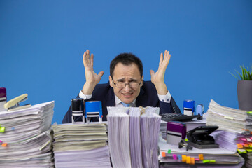 tired middle-aged stressed and overworked businessman wearing glasses yelling in office with huge stack of documents in giving up concept. overworking concept. isolated on blue overhour job