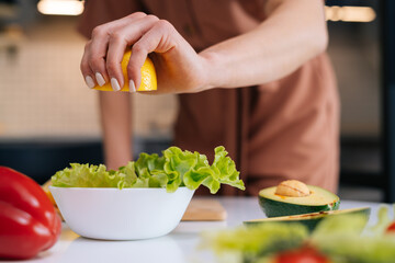 Close-up of women's hands squeezing juice from fresh yellow lemon into salad bowl with sliced vegetables. Red bell pepper, avocado, lemon on the table. Concept of healthy food lifestyle.