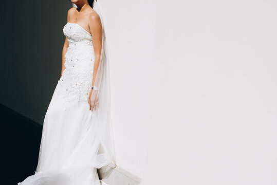 Bride In A White Dress With Long Veil Stands Against A White Wall. Light And Shadows. Beautiful Charming Bride In Wedding Luxurious Dress Posing Against A White Wall. No Face, Space For Text