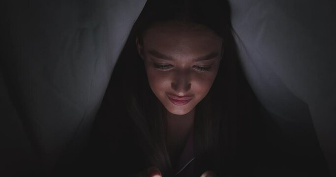 Young woman web surfing on smartphone, hiding under duvet in bed