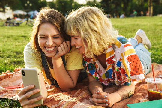 Image of joyful two women using cellphone while have picnic on grass